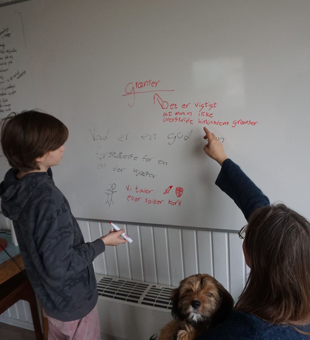 Et barn skriver på en whiteboardtavle, mens to voksne sidder ved siden af og kigger med.